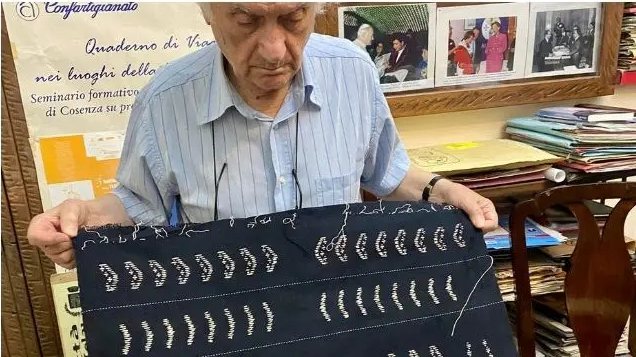 Un uomo anziano mostra un tessuto blu notte con decori chiari, in un ufficio con fotografie d'archivio alle pareti