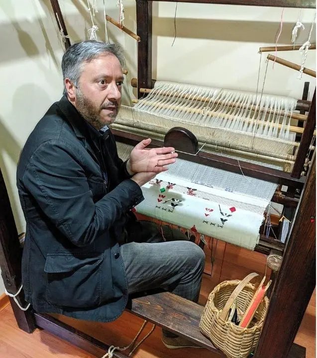 Un uomo seduto al telaio di legno tradizionale, con l'ordito teso e un tessuto bianco decorato in lavorazione